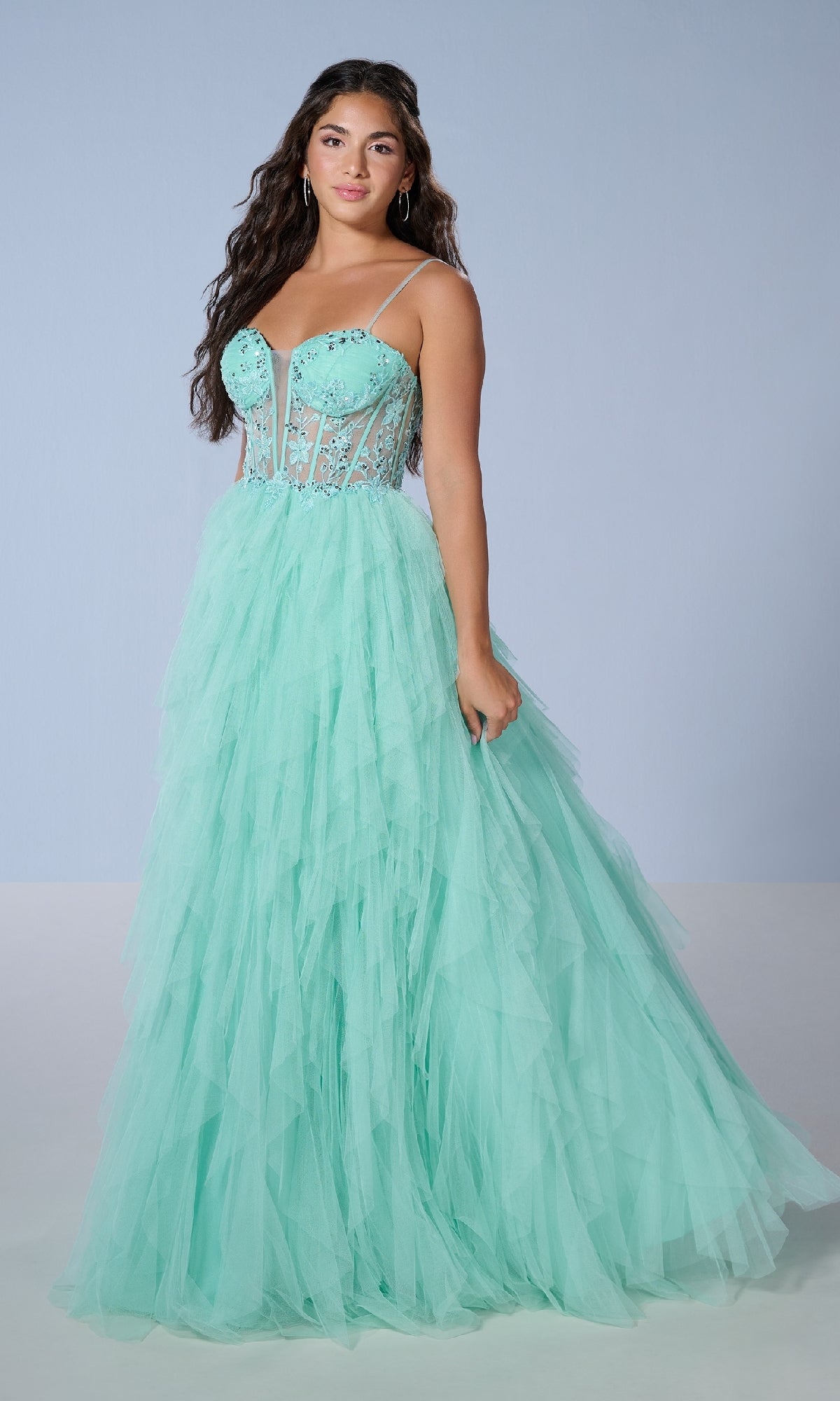 A woman wears the Long Prom Dress: Tiffany 16149, a light blue tulle gown with a corset-style bodice, thin straps, and a layered skirt with cascading ruffles, posed against a plain light background.